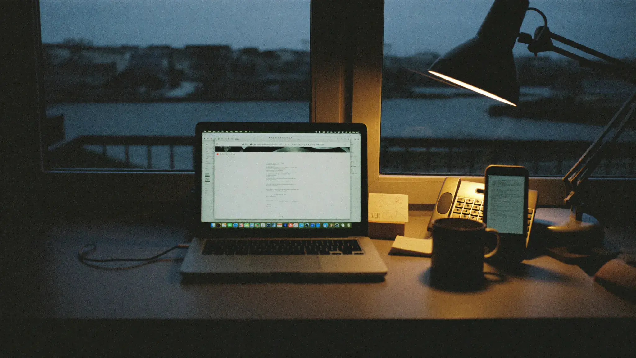 A tidy home office desk at night with a phone face-up beside a laptop