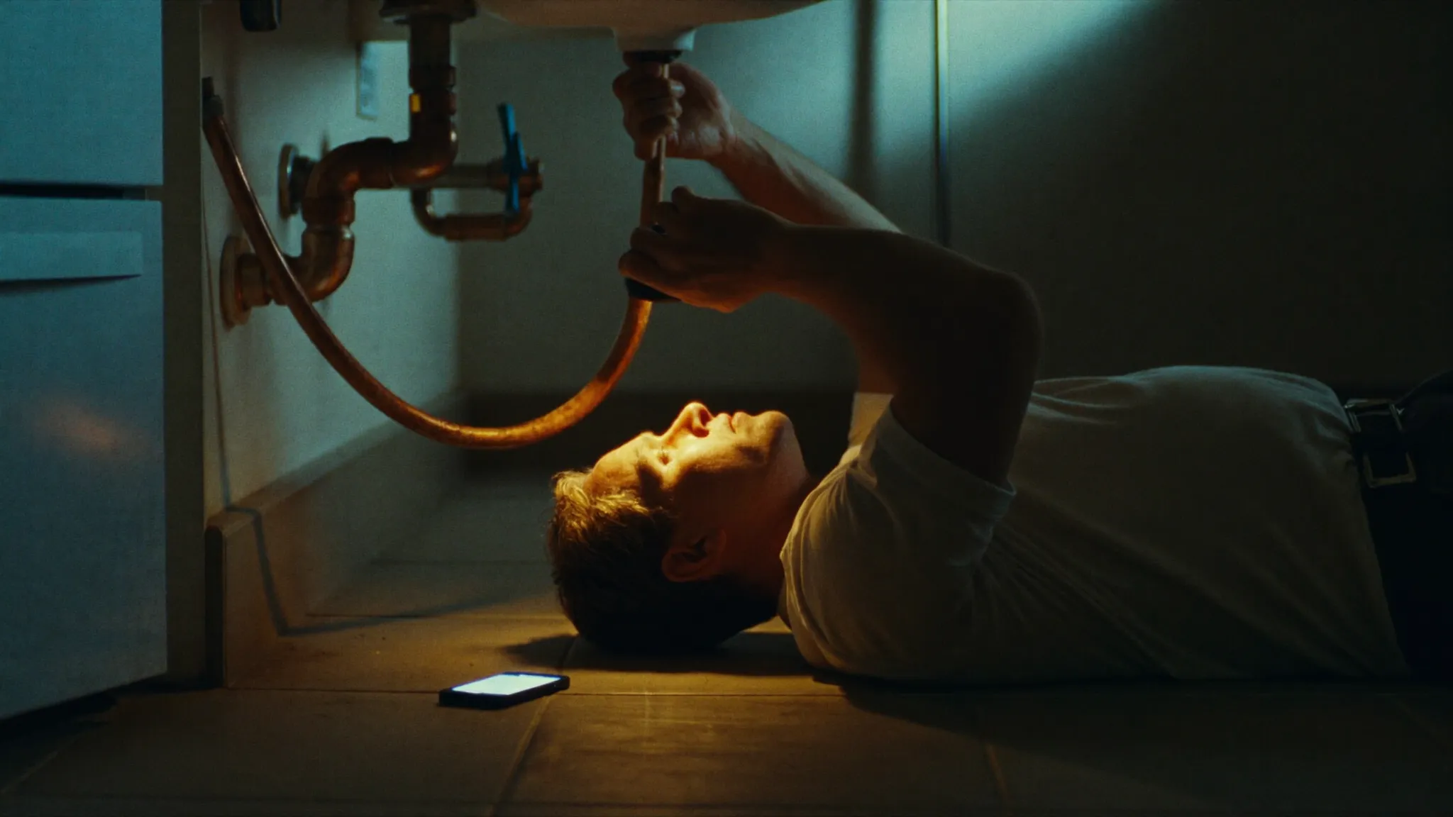 A plumber working under a sink by torchlight while his phone rings unanswered